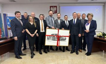 Grupowe zdjęcie przedstawiające 13 osób. Mężczyźni ustawieni w środku trzymią dużą oprawioną czarno-białą grafikę z szybkim samochodem bez dachu. Grafika przewiązana jest czerwoną wstążką. Zdjęcie wykonane jest w sali konwerencyjnej. po bokach częściowo widać stoły. Na ścianie widac zawieszkone godło Polski z lewej strony tablicę z częćiowo widocznym napisem narada roczna. Z prawej strony stoi uśmiechnięta kobieta z lewej strony obok mężczyzny również stoi kobieta.